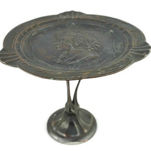 May include: A silver-toned metal serving tray with a raised design of a woman's face. The tray is on a stand with a decorative base.