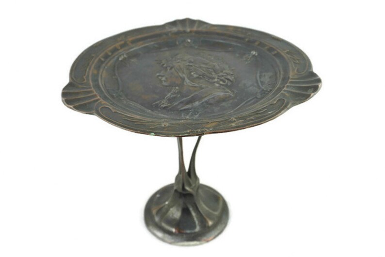 May include: A silver-toned metal serving tray with a raised design of a woman's face. The tray is on a stand with a decorative base.