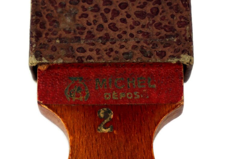 May include: A wooden razor strop with a red leather top and a brown leather backing. The strop has the text "MICHEL DEPOS." and the number "2" printed on it.