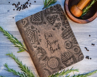 May include: A wooden recipe book with a brown cover and a design of various foods, including pizza, waffles, donuts, and french fries. The text "Our Recipe Book" is engraved on the cover.