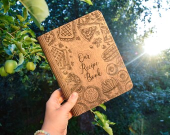 May include: A wooden recipe book with a brown cover and a design of various foods, including pizza, donuts, fries, and waffles. The text "Our Recipe Book" is engraved on the cover.