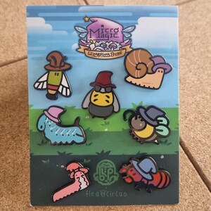 May include: A set of seven enamel pins featuring whimsical insect and animal characters wearing hats. The pins include a snail, bee, ant, and other creatures. The packaging has the text "Micro Magic Whispering Beast" and "Hea8Circus".
