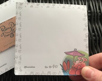 May include: A set of two blank notepads with a whimsical illustration of a pink creature under an umbrella. The notepads have a white background with a gray border of repeating rain cloud designs. The notepads are labeled "Rainy Day Thoughts" and "@brainblips".