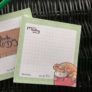 Könnte beinhalten: Zwei grüne und weiße Notizblöcke mit einem Gittermuster. Der obere Block hat ein braunes Etikett mit dem Text "Apple a Day Handmade Memo" und der untere Block hat ein Cartoon-Hamster, der einen Apfel isst. Beide Blöcke haben das Wort "Montag" darauf geschrieben.