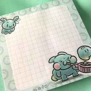 May include: A white notepad with a grid pattern and a light blue border. The notepad has three cartoon characters on it. One character is a light blue bunny with a pink nose and white wings. Another character is a light blue bunny with a pink nose and a pink pipe. The third character is a light blue bunny with a pink nose and a light blue halo. The notepad has the words "Etsy", "Instagram", and "Twitter" written on the bottom.