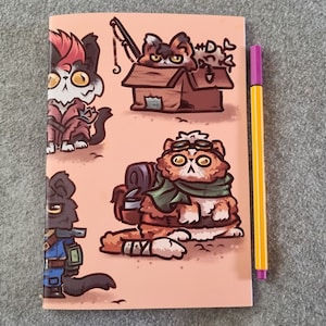 Könnte beinhalten: Ein Notizbuch mit rosa Einband mit Illustrationen von Cartoon-Katzen in verschiedenen Outfits. Ein lila-gelber Stift liegt neben dem Notizbuch. Die Katzen sind mit verschiedenen Accessoires dargestellt, darunter Brillen, Rucksäcke und Angelausrüstung.
