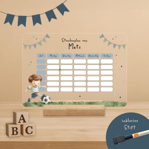 Puede incluir: Una pizarra acrílica transparente con horario semanal con el texto "Stundenplan von Mats" y una ilustración de fútbol. Incluye una base de madera y un rotulador. La pizarra tiene una cuadrícula para programar y está decorada con banderines azules.