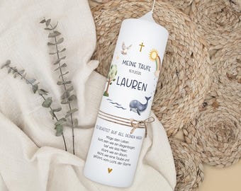 Op de afbeelding: Witte pilaar kaars met een decoratief ontwerp. De kaars heeft de tekst "MEINE TAUFE LAUREN 15.11.2025" met illustraties van een regenboog, walvis, boom en duif. Een bruine touwstrik is om de kaars gebonden.