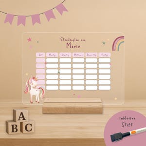 Puede incluir: Una pizarra acrílica transparente con horario semanal y base de madera. La pizarra presenta un gráfico de unicornio rosa y blanco, un arcoíris y el texto "Stundenplan von Marie". Incluye un rotulador de pizarra blanca negro.