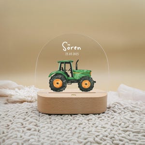 Può includere: Una luce notturna in acrilico trasparente con una base in legno. Un trattore verde con una ruota gialla è stampato sulla luce notturna. Il testo "Sören 23.01.2023" è stampato sull'acrilico.