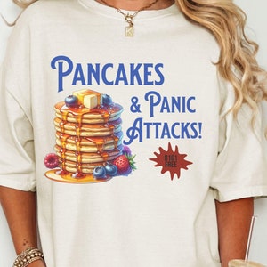 May include: Cream-colored t-shirt with the text "Pancakes & Panic Attacks!" in blue. A stack of pancakes with butter, blueberries, and syrup is illustrated below the text. A red burst graphic reads "B1G1 FREE".