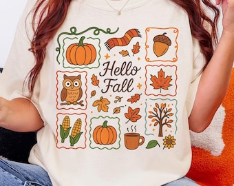 May include: Ivory-colored t-shirt featuring a colorful "Hello Fall" graphic. The design includes pumpkins, leaves, an owl, and a scarf. The shirt is a comfortable, casual style, perfect for autumn.