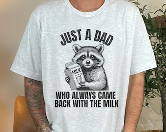 May include: Light gray t-shirt featuring a detailed illustration of a raccoon holding a milk carton. The text reads "JUST A DAD WHO ALWAYS CAME BACK WITH THE MILK" in a bold, black font. The shirt is a casual wear item.