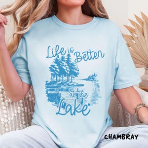 Puede incluir: Una camiseta azul claro con el texto "Life is Better at the Lake" en letras azules. El dise&ntilde;o presenta una escena de lago con &aacute;rboles y un muelle.
