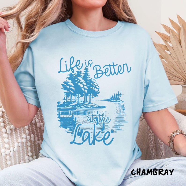 Puede incluir: Una camiseta azul claro con el texto "Life is Better at the Lake" en letras azules. El dise&ntilde;o presenta una escena de lago con &aacute;rboles y un muelle.