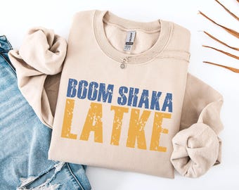 May include: A sand-colored sweatshirt with the words "BOOM SHAKA LATKE" printed in blue and yellow. The sweatshirt is folded and placed on a white surface next to a pair of blue jeans.