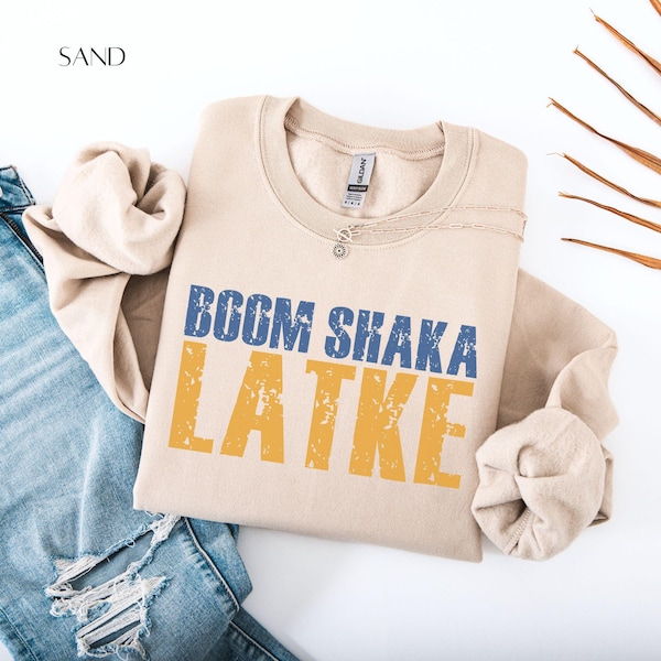 May include: A sand-colored sweatshirt with the words "BOOM SHAKA LATKE" printed in blue and yellow. The sweatshirt is folded and placed on a white surface next to a pair of blue jeans.