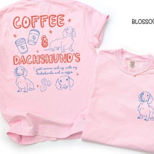 May include: Two light pink t-shirts. One has a dachshund graphic on the pocket. The other has a dachshund graphic, coffee cups, and the words "Coffee & Dachshund's" in red and blue. The text "I just wanna curl up with my dachshunds and a coffee" is also present.