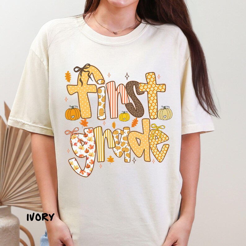 Op de afbeelding: Ivoor kleurig T-shirt met de tekst "First Grade" in een herfstthema ontwerp. De tekst bevat patronen van pompoenen, bladeren en strepen in tinten oranje, bruin en geel. Het shirt heeft korte mouwen en een ronde hals.