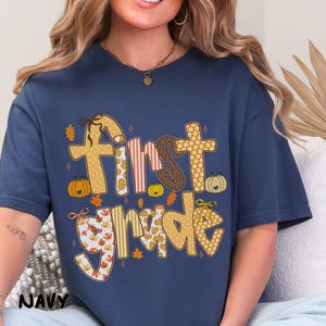 Op de afbeelding: Marineblauw T-shirt met de tekst "First Grade" in een herfstthema-ontwerp. De tekst bevat pompoenen, bladeren en verschillende patronen in oranje, bruin en geel. Het shirt is een comfortabele, casual stijl.