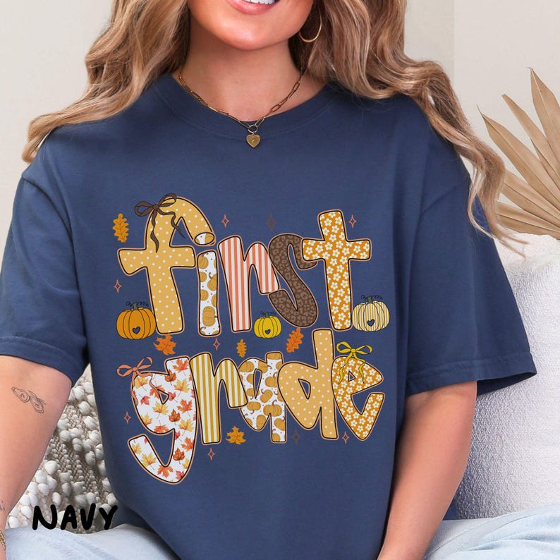 Op de afbeelding: Marineblauw T-shirt met de tekst "First Grade" in een herfstthema-ontwerp. De tekst bevat pompoenen, bladeren en verschillende patronen in oranje, bruin en geel. Het shirt is een comfortabele, casual stijl.