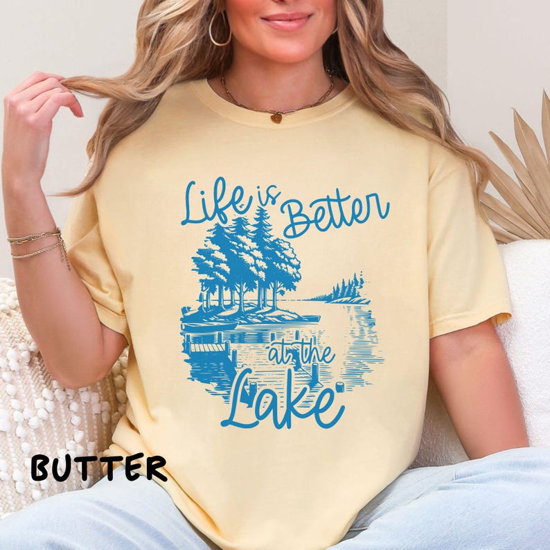 Puede incluir: Una camiseta amarilla con un gr&aacute;fico azul que dice "Life is Better at the Lake". El gr&aacute;fico presenta una escena de lago con &aacute;rboles y un bote.