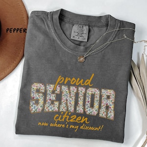 May include: A gray Comfort Colors t-shirt with the words "proud SENIOR citizen" in gold and floral print. The text "now where's my discount!" is below. A silver necklace with a pendant is on the shirt.