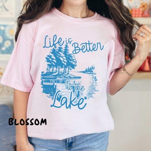 Puede incluir: Una camiseta rosa con un gr&aacute;fico azul que dice "Life is Better at the Lake". El gr&aacute;fico presenta una escena de lago con &aacute;rboles y un muelle.