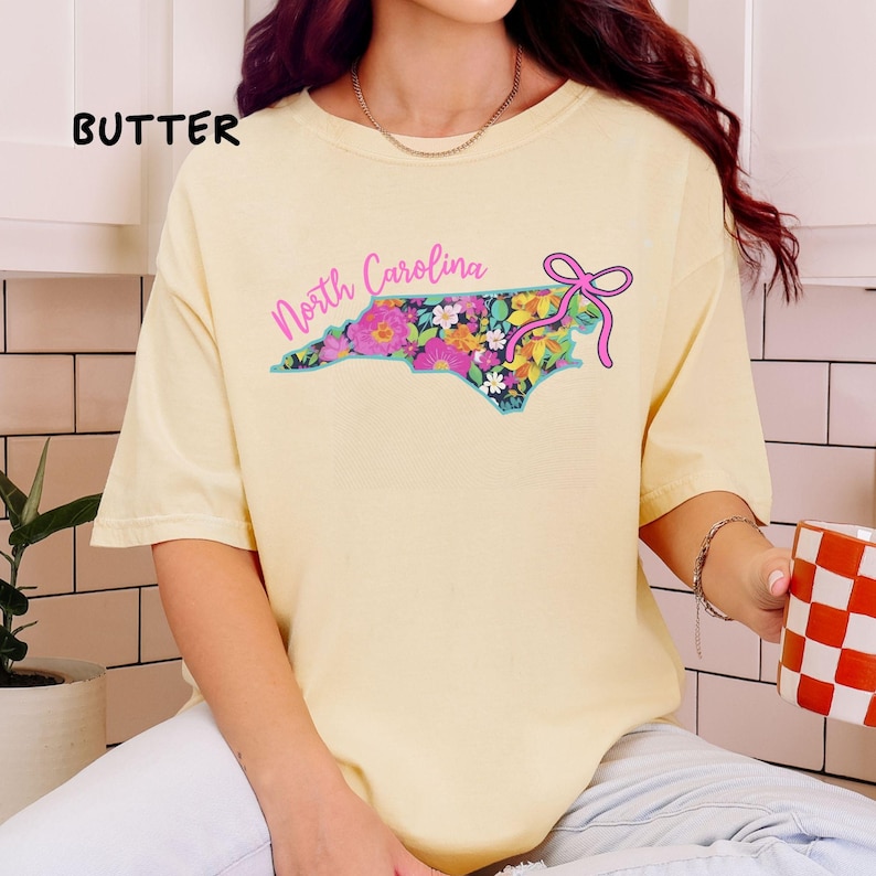 May include: A light yellow t-shirt with the word "BUTTER" in black block letters. The shirt features a floral design in the shape of North Carolina with the words "North Carolina" in pink script and a pink bow.