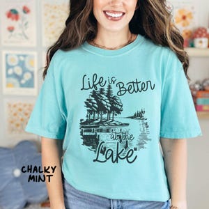 Puede incluir: Una camiseta azul turquesa con el texto "Life is Better at the Lake" en letras negras. El dise&ntilde;o presenta una silueta negra de un lago con &aacute;rboles y un muelle.