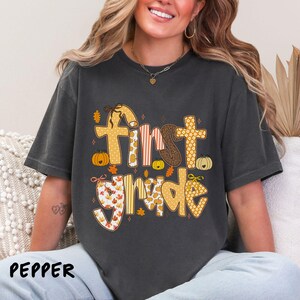 Op de afbeelding: Donkergrijs t-shirt met de woorden "First Grade" in een herfstthema ontwerp. De tekst heeft oranje, bruine en witte patronen, met pompoenen en bladeren. Het woord "PEPPER" staat linksonder.