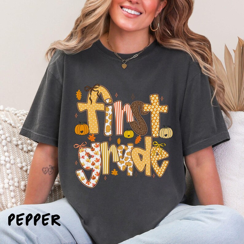 Op de afbeelding: Donkergrijs t-shirt met de woorden "First Grade" in een herfstthema ontwerp. De tekst heeft oranje, bruine en witte patronen, met pompoenen en bladeren. Het woord "PEPPER" staat linksonder.