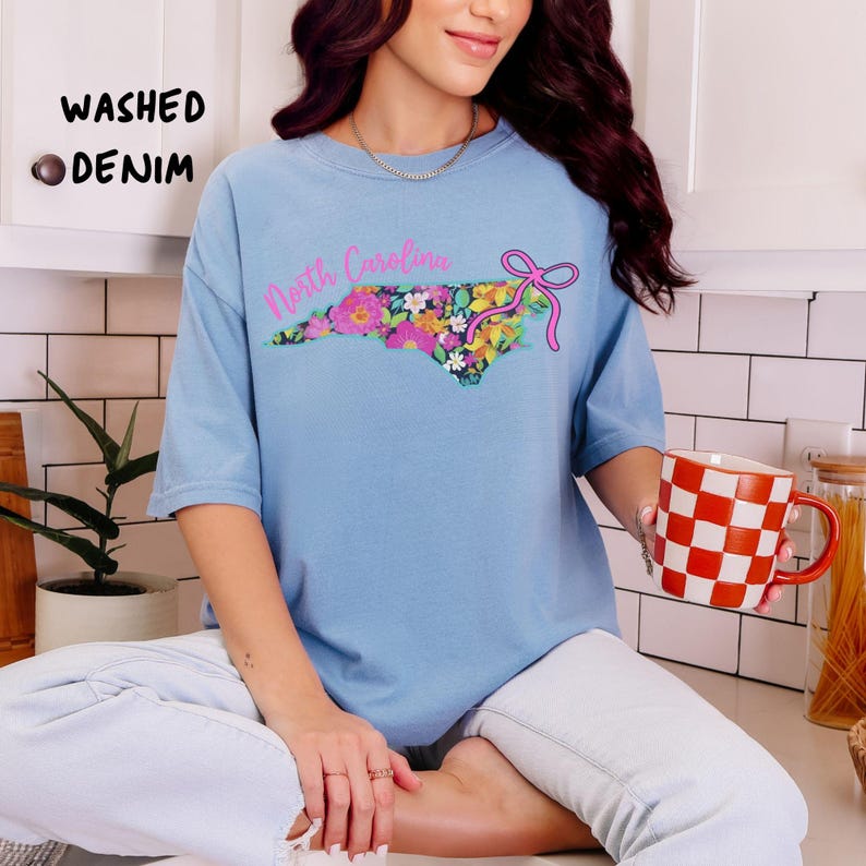 May include: Light blue t-shirt with a floral North Carolina state design and the words "North Carolina" in pink. The shirt also has the words "Washed Denim" printed on the upper left side.