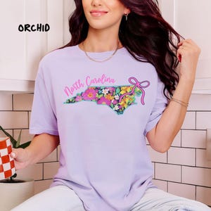 May include: A light purple t-shirt with the word "ORCHID" in black text. The shirt features a floral design in the shape of North Carolina, with the words "North Carolina" in pink script and a pink bow.