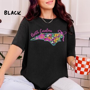 May include: Black t-shirt featuring a floral North Carolina state outline design. The state outline is filled with colorful flowers and the words "North Carolina" in pink script. A pink bow is on the right side of the state outline.
