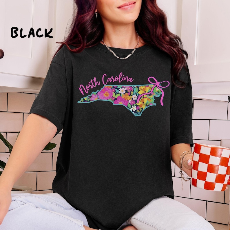 May include: Black t-shirt featuring a floral North Carolina state outline design. The state outline is filled with colorful flowers and the words "North Carolina" in pink script. A pink bow is on the right side of the state outline.