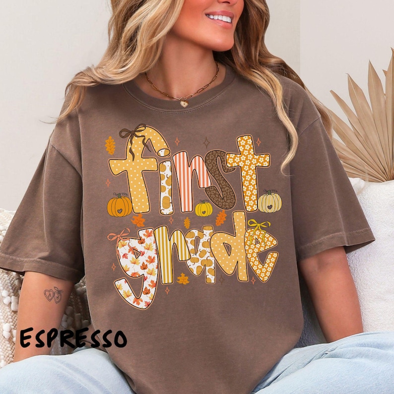 Op de afbeelding: Espresso-kleurig T-shirt met de woorden "First Grade" in een herfstthema-ontwerp. De tekst bevat pompoenen, bladeren en verschillende patronen in tinten geel, oranje en bruin. Het shirt heeft een relaxte pasvorm.
