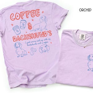 May include: Orchid-colored t-shirts with dachshund and coffee-themed graphics. One shirt features a large design on the back with text and illustrations, while the other has a small dachshund graphic on the front pocket.