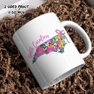 May include: White 11 oz mug featuring a floral design in the shape of North Carolina. The design includes pink, yellow, and blue flowers with a pink bow. The mug has the text "North Carolina" in a cursive font.