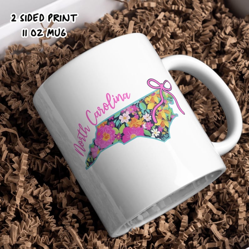 May include: White 11 oz mug featuring a floral design in the shape of North Carolina. The design includes pink, yellow, and blue flowers with a pink bow. The mug has the text "North Carolina" in a cursive font.