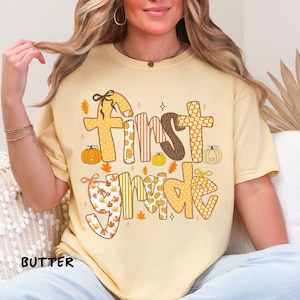 Op de afbeelding: Een lichtgeel T-shirt met de tekst "First Grade" in een herfstthema-ontwerp. De tekst bevat verschillende patronen, waaronder pompoenen, bladeren en bloemenprints. Het woord "BUTTER" staat linksonder.