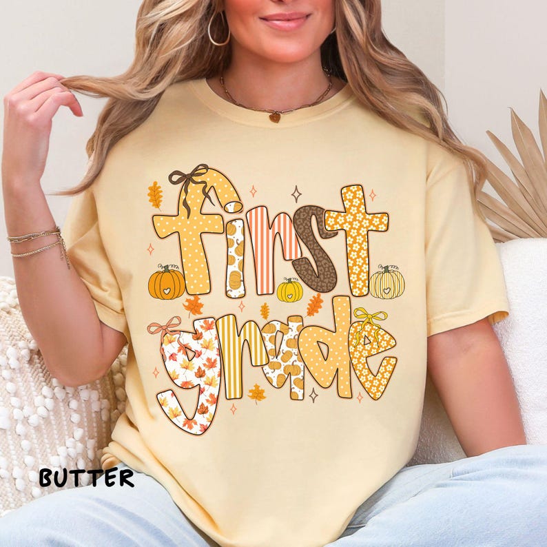 Op de afbeelding: Een lichtgeel T-shirt met de tekst "First Grade" in een herfstthema-ontwerp. De tekst bevat verschillende patronen, waaronder pompoenen, bladeren en bloemenprints. Het woord "BUTTER" staat linksonder.