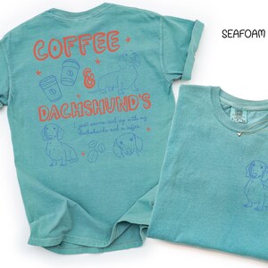 May include: Seafoam green t-shirts with dachshund and coffee-themed graphics. One shirt has a large design on the back with text and illustrations, while the other has a small dachshund graphic on the front pocket area. A gold necklace is draped on the second shirt.
