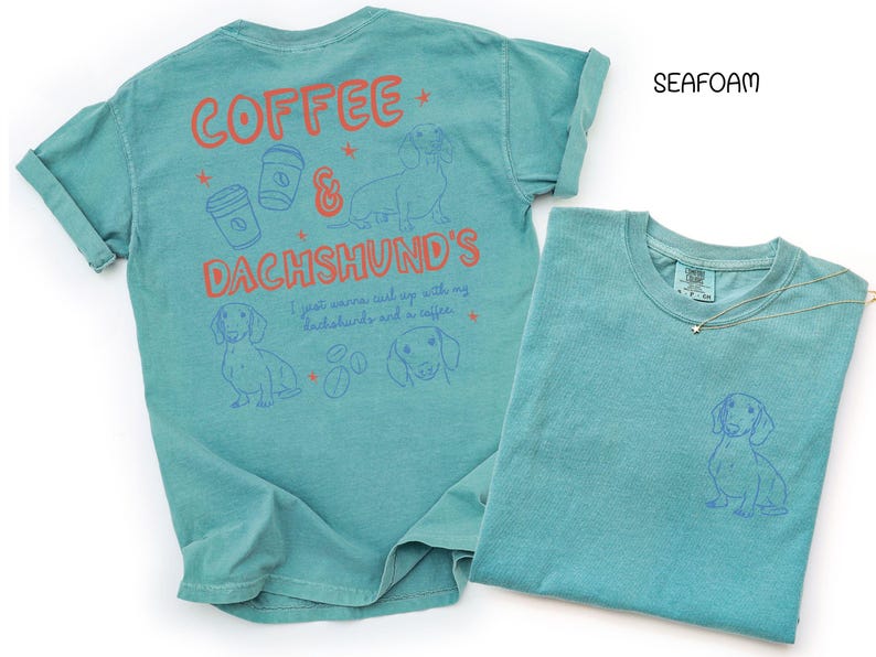 May include: Seafoam green t-shirts with dachshund and coffee-themed graphics. One shirt has a large design on the back with text and illustrations, while the other has a small dachshund graphic on the front pocket area. A gold necklace is draped on the second shirt.