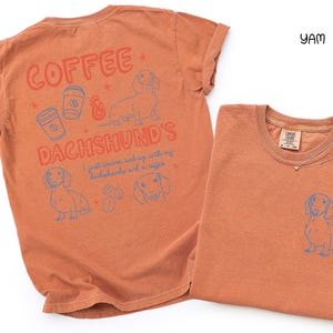 May include: Two orange-colored t-shirts. One shirt has the text "Coffee & Dachshund's" with illustrations of coffee cups and a dachshund. The other shirt has a small dachshund illustration on the front. The shirts are made of a soft material.