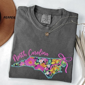 May include: A gray Comfort Colors t-shirt featuring a floral North Carolina state outline with a pink bow. The shirt has a crew neck and short sleeves. A gold pendant necklace is draped on the shirt.