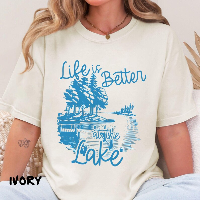 Puede incluir: Una camiseta blanca con un gr&aacute;fico azul que dice "Life is Better at the Lake". El gr&aacute;fico presenta una escena de lago con &aacute;rboles y un bote.