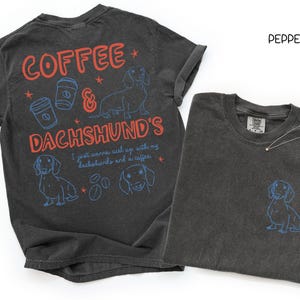 May include: Two dark gray t-shirts. One has the words "COFFEE & DACHSHUND'S" in red and blue, with dachshund and coffee cup graphics. The other shirt has a small dachshund graphic on the pocket.