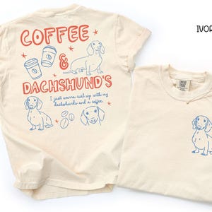 May include: Ivory-colored t-shirts with blue and red graphics. One shirt features a dachshund illustration on the front and a larger design on the back with the text "Coffee & Dachshund's". The other shirt has a dachshund illustration on the front.