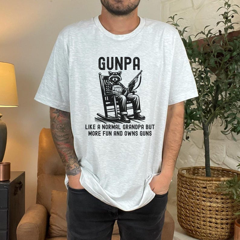May include: A gray t-shirt with a black and white illustration of a raccoon sitting in a rocking chair holding a gun. The text "Gunpa" is above the illustration and the text "Like a normal grandpa but more fun and owns guns" is below the illustration.